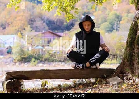 Uomo che pratica mudra yoga con l'acqua. L'uomo in abito nero pratica mudra yoga sulla panchina con vista sul fiume, sugli alberi autunnali e sul paesaggio montano Foto Stock