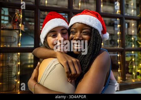 Amici felici e diversi che indossano cappelli di babbo natale, abbracciano e festeggiano insieme le vacanze Foto Stock