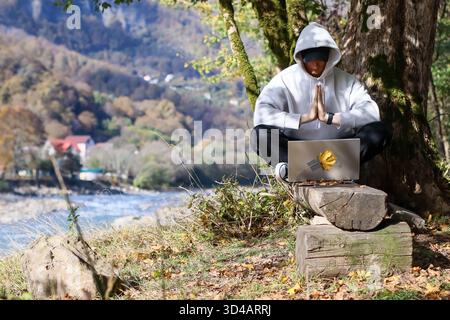 Persona in felpa con cappuccio con le mani in posa di preghiera con un portatile in natura. La figura con cappuccio si trova sotto l'albero con il laptop sul tronco, i palmi premuti insieme Foto Stock