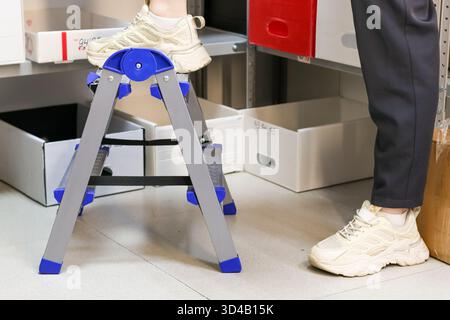 Piedi sulla scala a cavalletto vicino ai ripiani di stoccaggio nel punto di prelievo. Primo piano delle gambe della persona in piedi su una scala pieghevole accanto agli scaffali con scatole in consegna Foto Stock