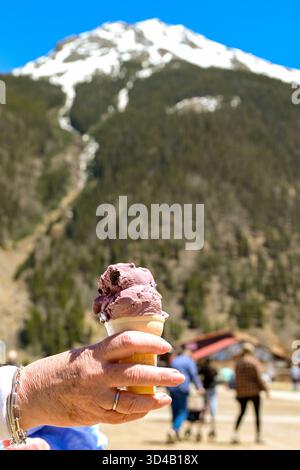 Vista ravvicinata di una persona in vacanza che tiene in mano un cono con gelato alla ciliegia con una montagna innevata sullo sfondo Foto Stock
