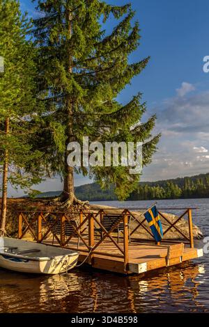 Barca a remi ormeggiata in un rustico molo in legno presso un tranquillo lago, caratterizzato da una vibrante bandiera svedese e circondato da lussureggianti alberi di pino verdi sotto un blu chiaro Foto Stock
