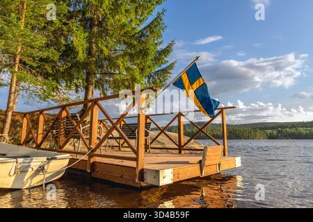 Bandiera svedese che sventola da un rustico molo in legno vicino a un lago tranquillo, una piccola barca legata a riva, circondata da verdi pini e da una lontana foresta sottostante Foto Stock