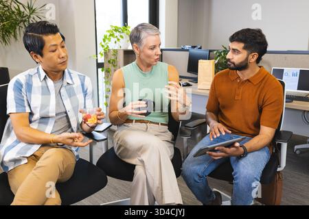 Colleghi diversi che discutono idee in un ufficio open space con tazza da caffè, tablet e contenitore di frutta Foto Stock