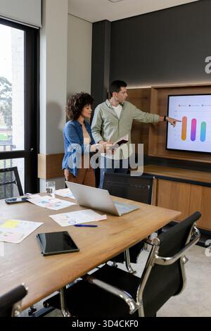 Revisione di diversi colleghi che utilizzano laptop e tablet nella sala conferenze, analisi dei grafici sullo schermo Foto Stock