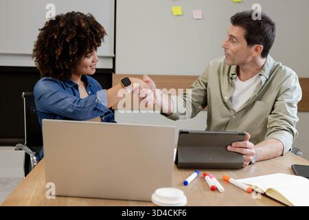 Diversi colleghi che stringono la mano sul tavolo da conferenza in ufficio, con laptop e tablet Foto Stock