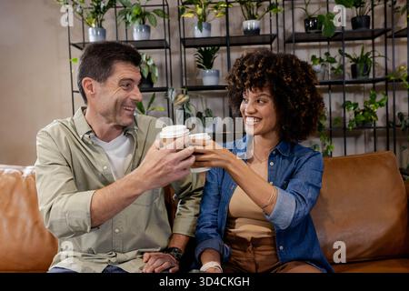 Amici diversi seduti su un divano in pelle nella sala moderna, con tazze da caffè usa e getta Foto Stock