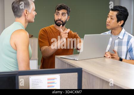 Brainstorming di colleghi diversi in piedi intorno alla scrivania in un ufficio moderno, utilizzando un computer portatile color argento Foto Stock