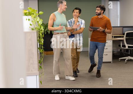 Camminare con colleghi diversi che discutono idee nel moderno corridoio dell'ufficio, tenendo in mano tablet e notebook Foto Stock