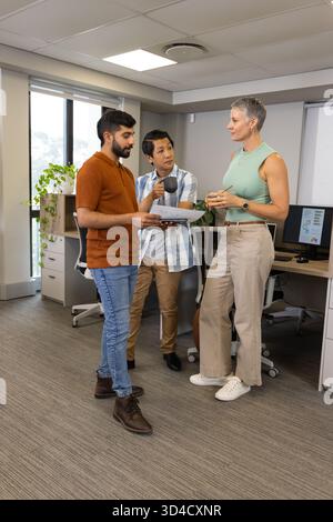 Diversi colleghi che discutono i dettagli del progetto in un ufficio aperto, con documenti stampati e tazze da caffè Foto Stock