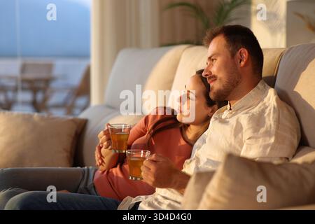 Coppia felice innamorata al tramonto che si abbraccia su un divano guardando lontano bevendo tè a casa Foto Stock