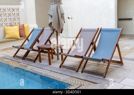 Scena a bordo piscina con tre poltrone in legno con cuscini in tessuto, un piccolo tavolo in legno e un ombrellone. Perfetto per il relax in piscina in un ambiente all'aperto sereno Foto Stock
