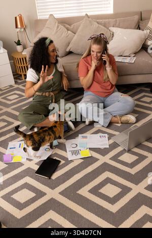 Collaborare con diverse amiche che lavorano sulle classifiche del tappeto da salotto, con un laptop aperto e un gatto Foto Stock