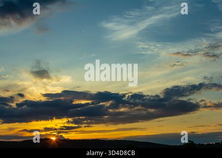 Uno splendido tramonto dipinge il cielo con sfumature di arancione e blu, mentre le nuvole si spostano pigramente sopra le colline ondulate creando una scena tranquilla nella campagna. Foto Stock