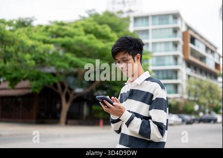 L'uomo asiatico che indossa gli occhiali sta guardando e scrivendo al suo smartphone mentre si trova nel parcheggio. In attesa o per riposarsi, rilassarsi all'aperto. Foto Stock
