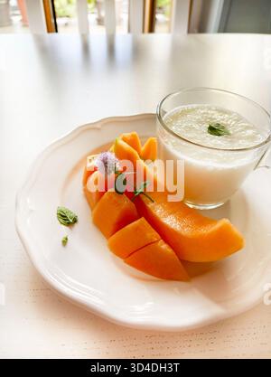 Fette di melone fresche e un bicchiere di yogurt o kefir su un piatto bianco, guarnite con foglie di menta. Un dessert o uno spuntino estivo. Concetto di cibo sano. Foto Stock