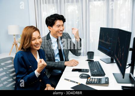 Partner commerciali asiatici sorridenti di successo che si alzano in piedi insieme in un lavoro di progetto redditizio nel database delle borse su pc che indossa una tuta formale a. Foto Stock