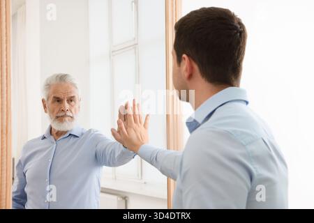 Giovane uomo che si vede allo specchio come un uomo anziano. Concetto di passato e futuro, flusso del tempo, età Foto Stock