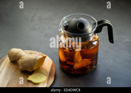 Tè caldo allo zenzero in una teiera di vetro con fette di zenzero fresche, su un tavolo da cucina grigio Foto Stock