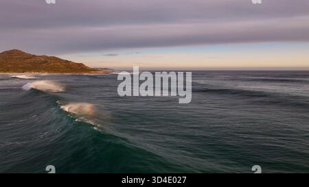 Vista aerea delle tumultuose onde oceaniche che si infrangono contro la costa frastagliata vicino a città del Capo, una maestosa danza del mare e della terra sotto un cielo sereno, C. Foto Stock