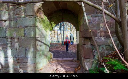 Vista dell'arco di pietra intemprata che incornicia un sentiero cosparso di foglie autunnali, che attira due figure in una scena boschiva dondolata dal sole, Haddington, Scotl Foto Stock