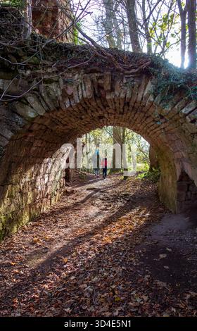 La vista di un arco di pietra intemperie incornicia figure che camminano attraverso un sentiero boschivo illuminato dal sole, foglie sparse sotto i piedi, una scena di fascino rustico, Haddi Foto Stock