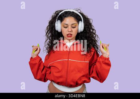 Giovane donna che ascolta la musica con le cuffie wireless a dondolo sullo sfondo viola Foto Stock