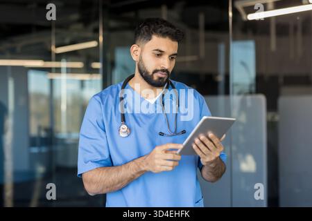 Medico indiano maschile in scrub blu con stetoscopio, focalizzato sulla revisione dei dati dei pazienti su una compressa in una clinica moderna e luminosa, simboleggiando la guarigione digitale Foto Stock