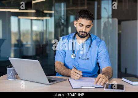 Giovane medico o infermiere maschile che indossa scrub e uno stetoscopio, seduto a una scrivania e compilando i documenti con un computer portatile e un telefono nelle vicinanze in un moderno cl Foto Stock