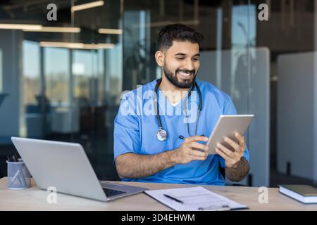 Medico indiano maschile o infermiere che indossa uniforme scrub e stetoscopio sorridente durante la revisione medica, informazioni su una compressa digitale in una clinica moderna Foto Stock