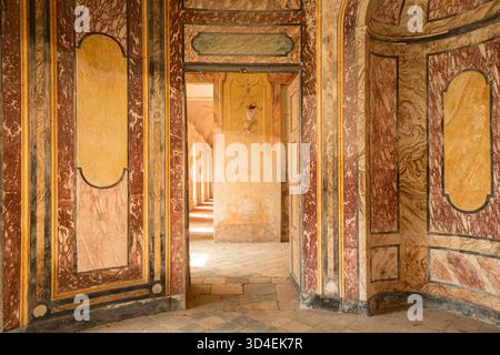 AGLIÈ, ITALIA? OTTOBRE 2025: Vista interna del castello agli è con pareti dipinte ornamentali, marmi invecchiati e porte che conducono ad un corridoio illuminato dal sole, simboleggiando il patrimonio e l'architettura reali italiani. Foto Stock