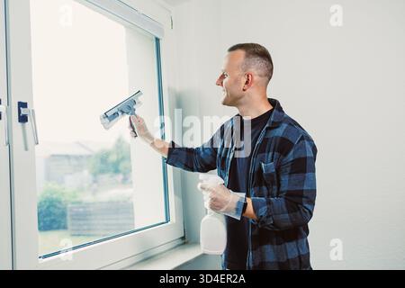 In una sala allegra piena di luce naturale, un uomo sta pulendo con piacere una finestra, utilizzando una bottiglia spray e un aspirapolvere per eliminare sporco e macchie, c Foto Stock