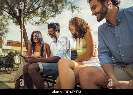 Quattro amici siedono su una panchina in un parco vivace in una giornata di sole, scambiandosi sorrisi e risate. Stanno condividendo qualcosa di divertente su uno smartphone, Foto Stock
