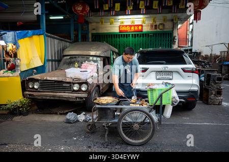 Giovane venditore che cucina cibo insano spazzatura su carrello mobile all'aperto in strada locale a Bangkok. Foto Stock