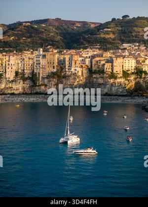 Veduta aerea di edifici baciati dal sole scendono giù dalle scogliere rocciose per incontrare il mare turchese, barche che rimbalzano dolcemente nel porto di Tropea, Calabria, Italia. Foto Stock