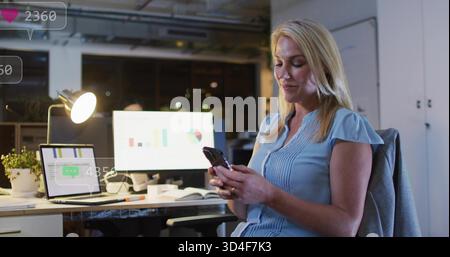 Tenere una donna bionda in una blusa azzurra che controlla il telefono nell'ufficio notturno con monitor e spazio fotocopie Foto Stock