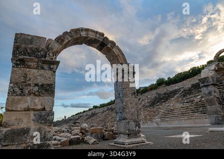 L'ingresso ad arco in pietra in rovina e le enormi rovine dell'antico teatro dietro di esso. Un sito storico e un patrimonio culturale. Foto Stock