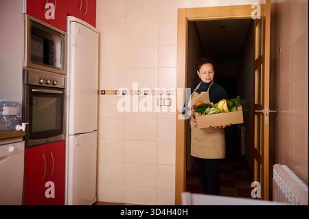 Una donna in un grembiule tiene una scatola di frutta e verdura fresca all'ingresso di una cucina luminosa, suggerendo cucina casalinga, negozi di alimentari e altro ancora Foto Stock
