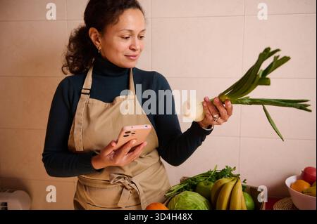 Una donna in un grembiule beige sta in una cucina, guardando il suo telefono mentre ispeziona un porro e una cipolla. Prodotti freschi e frutta sul bancone aggiungono un mar Foto Stock