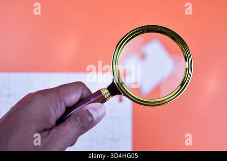 La lente d'ingrandimento sul puzzle bianco evidenzia il pezzo mancante su sfondo arancione, simboleggiando la ricerca mirata e avvicinandosi al completamento dell'attività strutturata Foto Stock