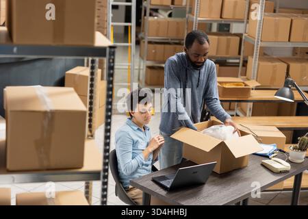 Team di dipendenti multietnici che controlla la logistica di consegna di una piccola azienda, sviluppando un marchio locale. Lo spazio del deposito industriale mostra il coordinamento logistico attivo e il monitoraggio dell'inventario. Foto Stock