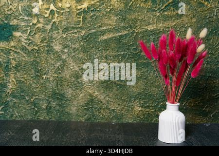 Un bouquet di fiori secchi rosa brillante si trova in un vaso bianco su un tavolo di legno scuro, creando un vibrante contrasto con la parete testurizzata verde dietro. Foto Stock