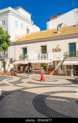 Lagos, Portogallo, 8 novembre 2025, Una donna in rosso attraversa una piazza illuminata dal sole con ciottoli bianchi e neri a onde nel centro della città. donna io Foto Stock
