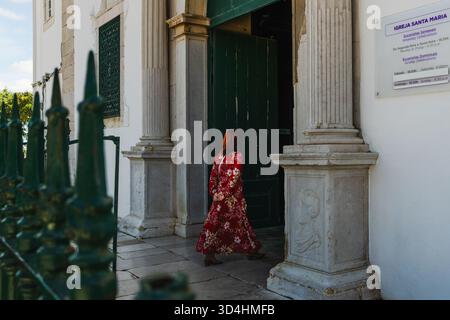 n: Lagos, Portogallo, 8 novembre 2025, donna con un abito floreale rosso entra nella storica Igreja Santa Maria attraverso la sua grande porta verde. Foto Stock