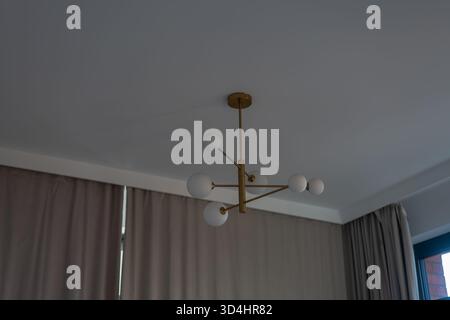 Un lampadario in ottone con globi bianchi è appeso a un soffitto bianco. Tende beige fiancheggiano le pareti, una cornice della finestra sulla destra suggerisce la luce della città a Danzica. Foto Stock