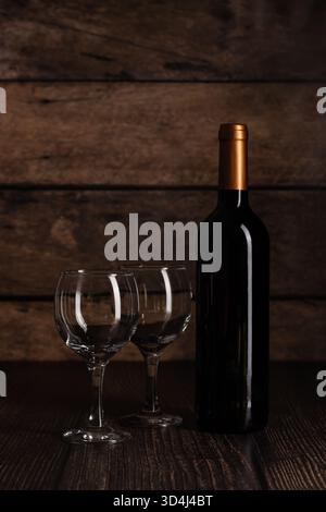 Una bottiglia di vino con tappo in bronzo si trova accanto a due bicchieri trasparenti su una superficie di legno scuro. La scena cattura un'atmosfera accogliente e invitante, perfetta Foto Stock