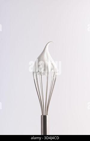 Una frusta di metallo ricoperta da picchi di panna montata soffice si erge in posizione verticale su un semplice sfondo. Una luce soffusa illumina la frusta, aggiungendo una sensazione di calore Foto Stock