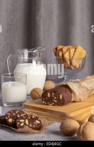 Il salame al cioccolato viene tagliato a fette e posto su un asse di legno accanto a un bicchiere di latte. Una caraffa di latte e una ciotola di biscotti sono dolci e invitanti Foto Stock
