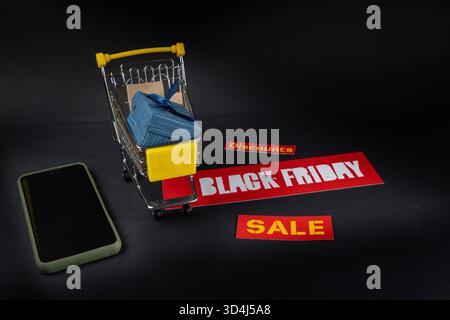 Il carrello della spesa in miniatura pieno di regali si trova su un cartello nero con i saldi del venerdì vicino allo smartphone su sfondo nero, concetto di shopping online Foto Stock