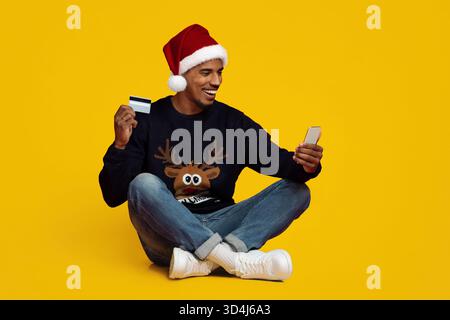 La persona che indossa il cappello Santa controlla il telefono mentre tiene in mano la carta di credito nel maglione festivo Foto Stock